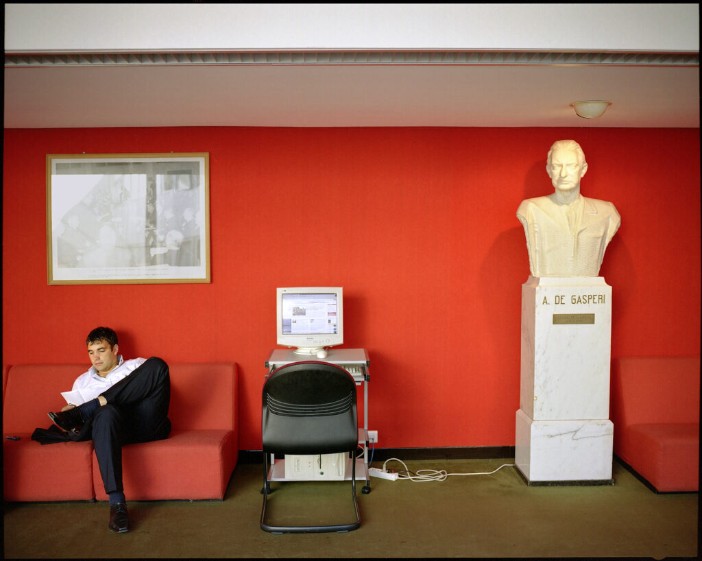 Un homme au travail est confortablement installé sur un canapé rouge au pied d'un buste de A de Gasperi, un ordinateur connecté à Internet est allumé au centre de l'image. 